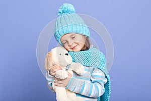 Studio shot of cute little girl dresses scarf, hat and warm shirt, holding her favorite toy, soft dog toy, child looking at her