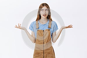 Studio shot of displeased bothered and questioned daughter in brown overalls shrugging turning look left feeling