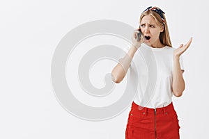 Studio shot of displeased moody girlfriend with blond hair frowning and compaining while talking on smartphone