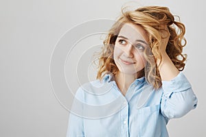 Studio shot of dreamy attractive woman with short curly hair looking aside with tender smile, touching haircut and