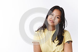 Studio shot of dreamy gentle young attractive asian female in yellow t-shirt tiltintg head tender and gazing at camera
