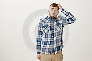 Studio shot of dumb handsome man in plaid shirt and falling glasses, scratching head and folding lips while saying wow