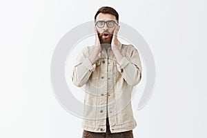 Studio shot of emotive shocked and worried male model with long beard in glasses holding hands on face looking nervous