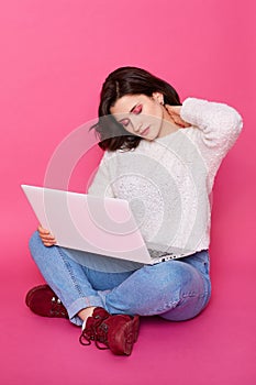 Studio shot of exhausted female with pain in neck, holding white lap top on her knee while sitting on floor, keeps eyes closed,