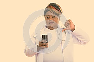 Studio shot of fat black African woman doctor holding glass of soda drink and giving thumb down