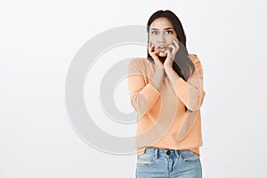 Studio shot of frightened intense attractive tanned female with dark hair, holding hands on face and dropping jaw while