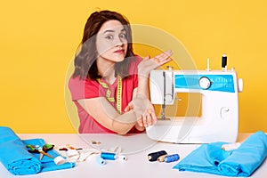 Studio shot of girl seamstress sitting at white desk with sewing machine, spreding hands, does not know how to switch it.