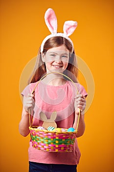 Studio Shot Of Girl Wearing Rabbit Ears And Holding Basket Of Easter Eggs Against Yellow Background