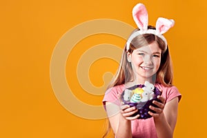 Studio Shot Of Girl Wearing Rabbit Ears And Holding Basket Of Easter Eggs Against Yellow Background