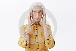 Studio shot of gloomy drained and upset old woman in trendy yellow coat holding hands on temples frowning from pain