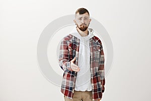 Studio shot of gloomy unconfident european guy with fair hair in trendy plaid shirt over hoodie, pulling hand towards