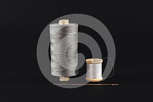 Studio shot of a grey and white thread reel, one big one small size, & a sewing needle isolated on black background. Spool (or