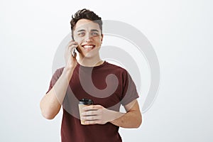 Studio shot of handsome cheerful guy, holding cup of coffee and talking on smartphone, smiling broadly, being satisfied