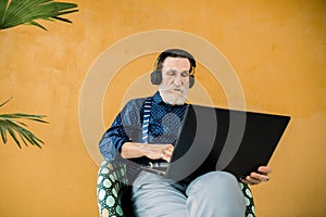 Studio shot of handsome concentrated senior man in stylish clothes, wearing earphones, sitting in the chair and using