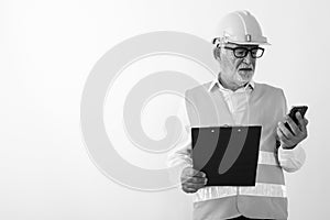 Studio shot of handsome senior bearded man construction worker using mobile phone while holding clipboard