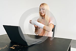 Studio shot of happy injured blonde young woman with broken arm wrapped in plaster bandage having video call online