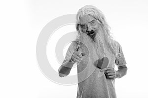 Studio shot of happy senior bearded man smiling and pointing at camera while holding heart ready for Valentine`s day