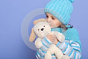 Studio shot of little charming girl wearing casually hugging and kissing her soft dog toy, posing in studio against blue wall,