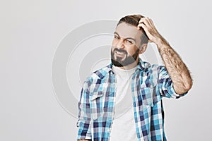 Studio shot of mad bearded male roughly scretching his head and expressing confusion, over gray background. Husband has