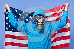 Studio shot of man ecologist holding USA flag behind him, wearing uniform and gas mask, posing isolated over blue background,