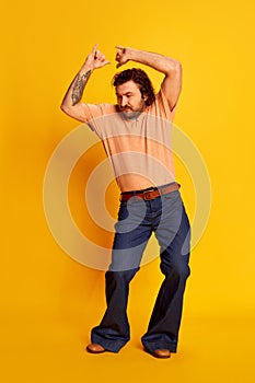 Studio shot of mature friendly bearded hairy man, hippie in stylish sunglasses and flared jeans dancing over yellow