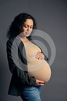 Studio shot multi-ethnic confident pregnant in beige bodysuit and stylish blazer, stroking her belly on gray background