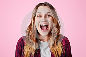 Studio shot of overjoyed happy female laughs at good joke, keeps mouth widely opened, being excited, isolated over pink studio bac