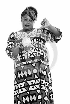 Studio shot of overweight African woman standing and holding piggy bank while giving thumb down