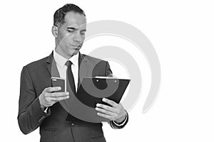 Mature handsome Persian businessman reading clipboard while holding mobile phone