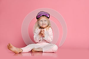 Studio shot of pleased beautiful young woman posing in eyemask. Cheerful Little cute girl in pajamas sits on the floor on pink photo