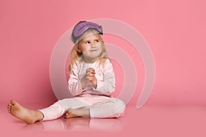 Studio shot of pleased beautiful young woman posing in eyemask. Cheerful Little cute girl in pajamas sits on the floor on pink