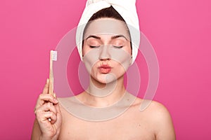 Studio shot of pleased female with eyes shut, pouts lips, wears white towel on head, woman holding toothbrush in hand, isolated