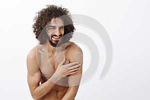 Studio shot of positive carefree handsome guy with beard and curly hair, bending while laughing out loud, touching chest