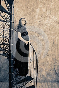 Studio shot of a pregnant woman standing on the step of a vintage spiral staircase, black and white