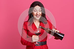 Studio shot of pretty young woman wearing red dresswith black belt, lady with long dark hair holding wine bottle and glass, girl
