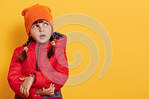 Studio shot of puzzled surprised Caucasian little girl looks surprisingly aside, has concerned expression, wears red jacket and