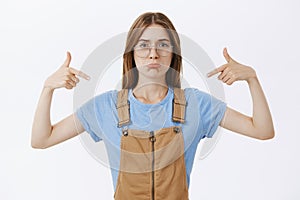 Studio shot of sad and gloomy unconfident female in glasses and brown dungarees pouting from sadness pointing at