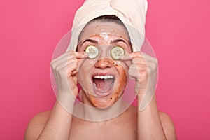 Studio shot of satisfaited woman applies slices of cucumber on her eyes and having scrabmask on face, has happy expresion, opens