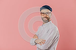 Studio shot of self assured bearded man with blue eyes, wears stylish hat and white shirt, keeps arms folded, models over pink