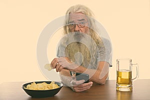 Studio shot of senior bearded man holding mobile phone while eating bowl of potato chips with glass of beer on wooden