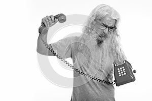 Studio shot of senior bearded man looking angry and shouting while holding old telephone