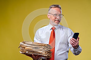 Studio shot of senior businessman looking at phone holding pile of papers. Yellow background