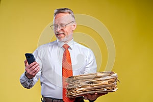 Studio shot of senior businessman looking at phone holding pile of papers. Yellow background