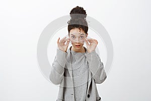 Studio shot of shocked disbelieving attractive european girl with curly hair combed in bun, taking off glasses, looking
