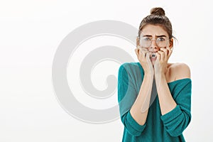 Studio shot of shocked and scared young woman in transparent glasses and loose sweater, frowning, biting fingernails