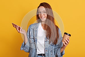 Studio shot of smiling young woman holding mobile phone and drinking take away coffee, surfing interner or chatting with friends,