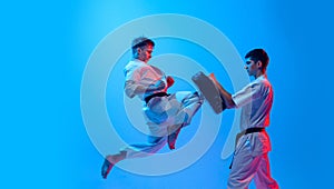 Studio shot of sports training of two karatedo fighters in doboks isolated on blue background in neon. Concept of combat
