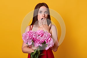 Studio shot of surprised young woman covering her opened mouth with hber palm, looks in amazement, holds bunch of pink peonies,