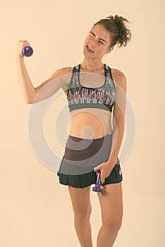 Studio shot of thoughtful teenage girl holding dumbbells while winking and sticking tongue out ready for sports and