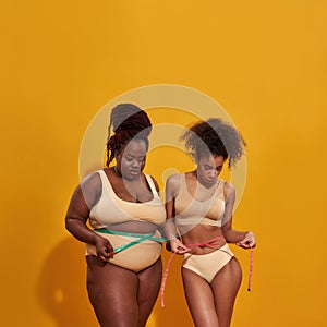 Studio shot of two african american women with different body weight measuring their waist using metric tape measure